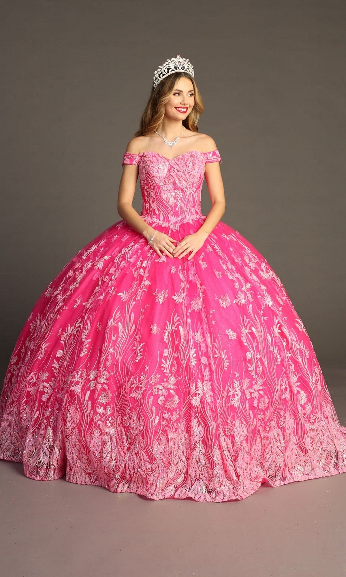 A woman wearing the Quinceanera Dress CHF3100 by Chicas—a pink, off-the-shoulder tulle ballgown adorned with white floral patterns—paired with a tiara and necklace, standing against a plain background.